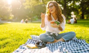 A smiling woman sits on a picnic blanket in a sunny park, eating a healthy bowl of salad. The image represents balanced eating and joyful lifestyle habits that complement Mounjaro (tirzepatide) weight loss treatment.