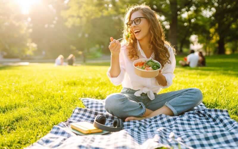 A smiling woman sits on a picnic blanket in a sunny park, eating a healthy bowl of salad. The image represents balanced eating and joyful lifestyle habits that complement Mounjaro (tirzepatide) weight loss treatment.