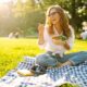 A smiling woman sits on a picnic blanket in a sunny park, eating a healthy bowl of salad. The image represents balanced eating and joyful lifestyle habits that complement Mounjaro (tirzepatide) weight loss treatment.
