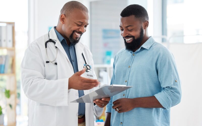 Smiling doctor discusses a treatment plan with a patient while reviewing a medical chart together in a bright clinic — representing supportive healthcare guidance and transparent discussion about medication costs.