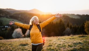 A joyful woman stands outdoors at sunset with her arms outstretched, wearing a yellow jacket and backpack, symbolizing energy, vitality, and rejuvenation — themes linked to the restorative benefits of NAD+ IV therapy and vitamin infusions.