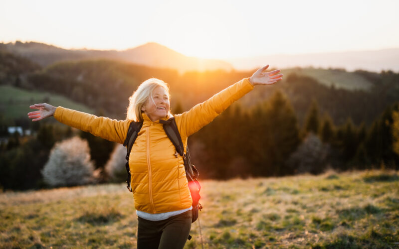 A joyful woman stands outdoors at sunset with her arms outstretched, wearing a yellow jacket and backpack, symbolizing energy, vitality, and rejuvenation — themes linked to the restorative benefits of NAD+ IV therapy and vitamin infusions.
