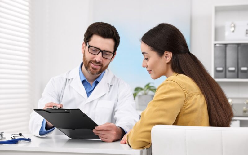 Smiling male doctor showing medical information on a clipboard to a female patient during a consultation, representing professional guidance on oral semaglutide (Rybelsus) dosing and safe weight management.