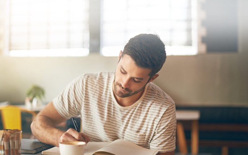Man sitting at a café writing in a notebook with a thoughtful expression, symbolizing self-reflection and emotional adjustment during a weight-loss journey with GLP-1 medications.