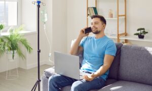 A relaxed, happy man sitting on a couch at home with an IV drip stand beside him, talking on the phone and using a laptop while receiving IV therapy.
