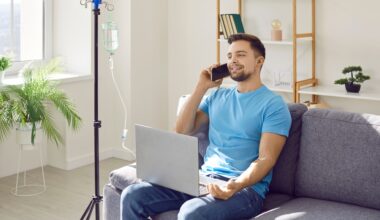 A relaxed, happy man sitting on a couch at home with an IV drip stand beside him, talking on the phone and using a laptop while receiving IV therapy.