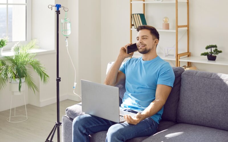A relaxed, happy man sitting on a couch at home with an IV drip stand beside him, talking on the phone and using a laptop while receiving IV therapy.