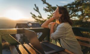 A happy woman sitting outdoors at a table with her laptop open, enjoying the fresh morning air while tracking her weight loss progress and lifestyle changes on Mounjaro (tirzepatide).
