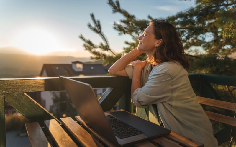 A happy woman sitting outdoors at a table with her laptop open, enjoying the fresh morning air while tracking her weight loss progress and lifestyle changes on Mounjaro (tirzepatide).