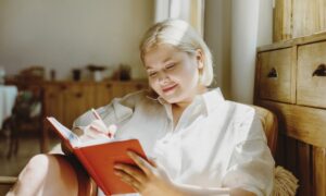 A woman sitting comfortably in a cozy, sunlit room writes in a red journal, appearing calm and reflective — symbolizing goal setting, self-awareness, and mindful tracking during her GLP-1 weight loss journey.