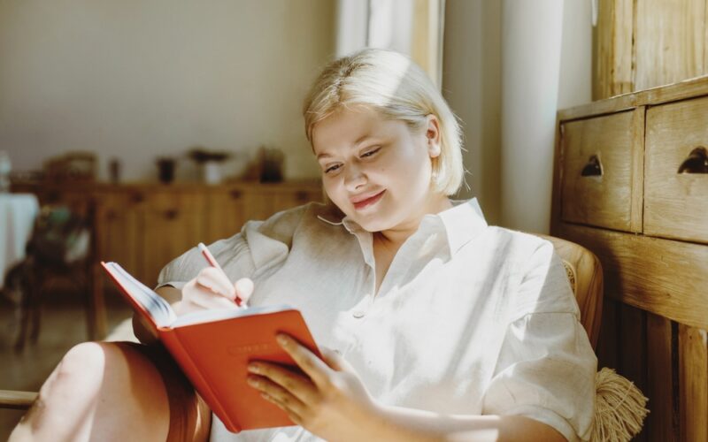 A woman sitting comfortably in a cozy, sunlit room writes in a red journal, appearing calm and reflective — symbolizing goal setting, self-awareness, and mindful tracking during her GLP-1 weight loss journey.
