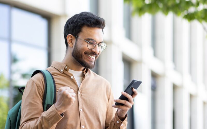 A smiling man outdoors looks excited while checking his phone, celebrating good news — representing successfully accessing a Zepbound discount or savings card.