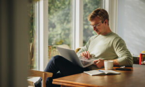 A man sits at a wooden table by a bright window, focused on his laptop with a notebook, coffee, and phone nearby—representing researching semaglutide prescriptions and taking proactive steps for weight management.