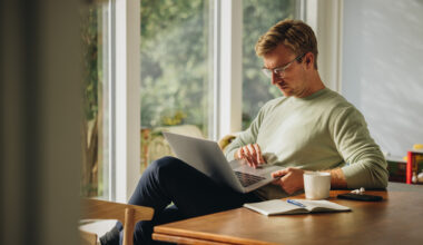 A man sits at a wooden table by a bright window, focused on his laptop with a notebook, coffee, and phone nearby—representing researching semaglutide prescriptions and taking proactive steps for weight management.