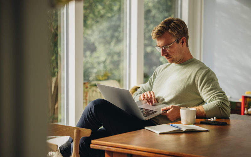 A man sits at a wooden table by a bright window, focused on his laptop with a notebook, coffee, and phone nearby—representing researching semaglutide prescriptions and taking proactive steps for weight management.