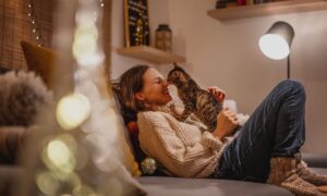 A woman relaxes on a cozy couch in a warmly lit living room, smiling as her cat gently touches noses with her — capturing the emotional comfort, bonding, and companionship central to life with an emotional support animal.