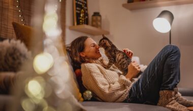 A woman relaxes on a cozy couch in a warmly lit living room, smiling as her cat gently touches noses with her — capturing the emotional comfort, bonding, and companionship central to life with an emotional support animal.