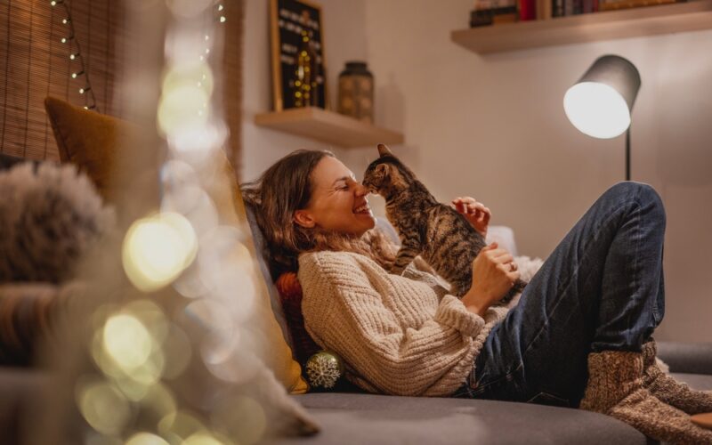 A woman relaxes on a cozy couch in a warmly lit living room, smiling as her cat gently touches noses with her — capturing the emotional comfort, bonding, and companionship central to life with an emotional support animal.