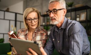 Older couple sitting together on a couch, reviewing medication information on a tablet while the woman holds a prescription pill bottle.