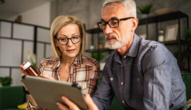 Older couple sitting together on a couch, reviewing medication information on a tablet while the woman holds a prescription pill bottle.