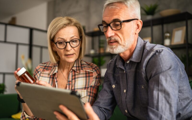 Older couple sitting together on a couch, reviewing medication information on a tablet while the woman holds a prescription pill bottle.