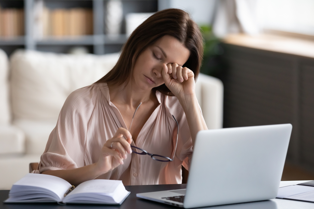 Tired woman sitting at a computer, resting her head on her hand, looking fatigued and overwhelmed, representing chronic fatigue syndrome and the potential role of B12 injections.