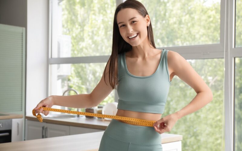 Smiling woman measuring her waist with a tape measure at home, representing hormone-driven weight loss, improved metabolism, and progress with GLP-1 medications.