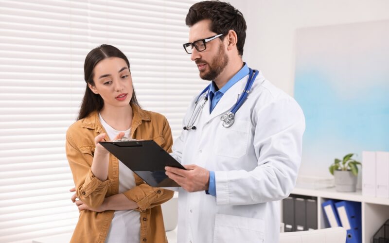 A doctor holding a clipboard explains a medication dosing plan to a woman in a clinical office, both reviewing information together.