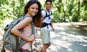 Two young adults hiking on a forest trail, smiling and wearing backpacks, enjoying an active outdoor lifestyle due to Peptide Therapy.