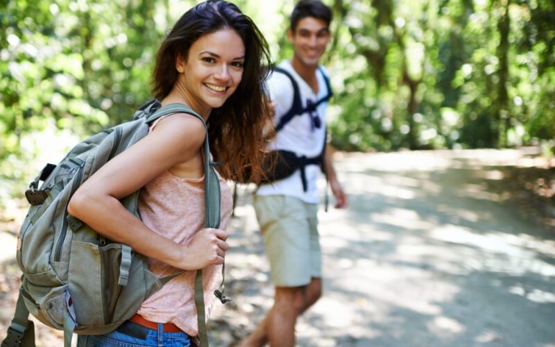 Two young adults hiking on a forest trail, smiling and wearing backpacks, enjoying an active outdoor lifestyle due to Peptide Therapy.