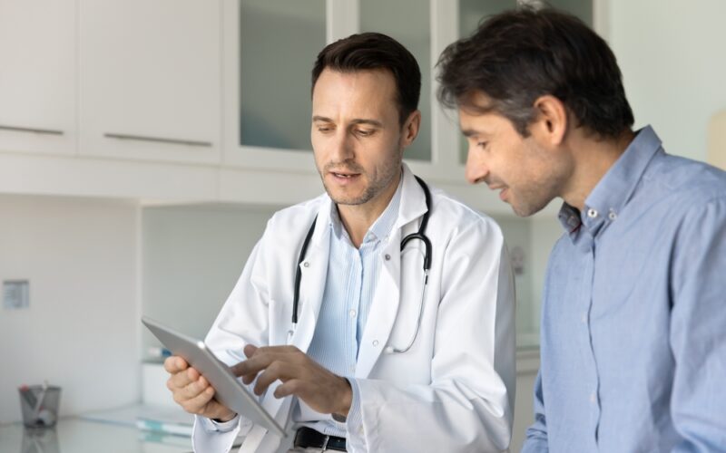 Male doctor showing a male patient information on an iPad, discussing hormonal health, testosterone levels, thyroid function, and treatment options for hormonal imbalance.