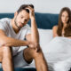 A man sitting on the edge of a bed looking concerned while a woman sits in the background, suggesting relationship or health-related stress.