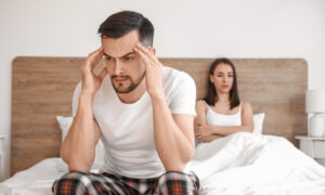 Man sitting on the edge of a bed holding his head while his partner sits behind him, illustrating stress, relationship strain, and common sexual health concerns such as erectile dysfunction and when to see a doctor.