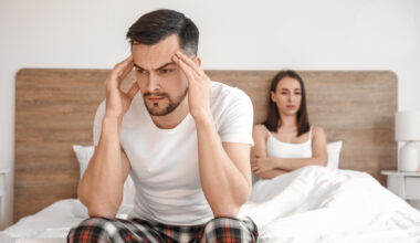 Man sitting on the edge of a bed holding his head while his partner sits behind him, illustrating stress, relationship strain, and common sexual health concerns such as erectile dysfunction and when to see a doctor.