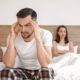 Man sitting on the edge of a bed holding his head while his partner sits behind him, illustrating stress, relationship strain, and common sexual health concerns such as erectile dysfunction and when to see a doctor.